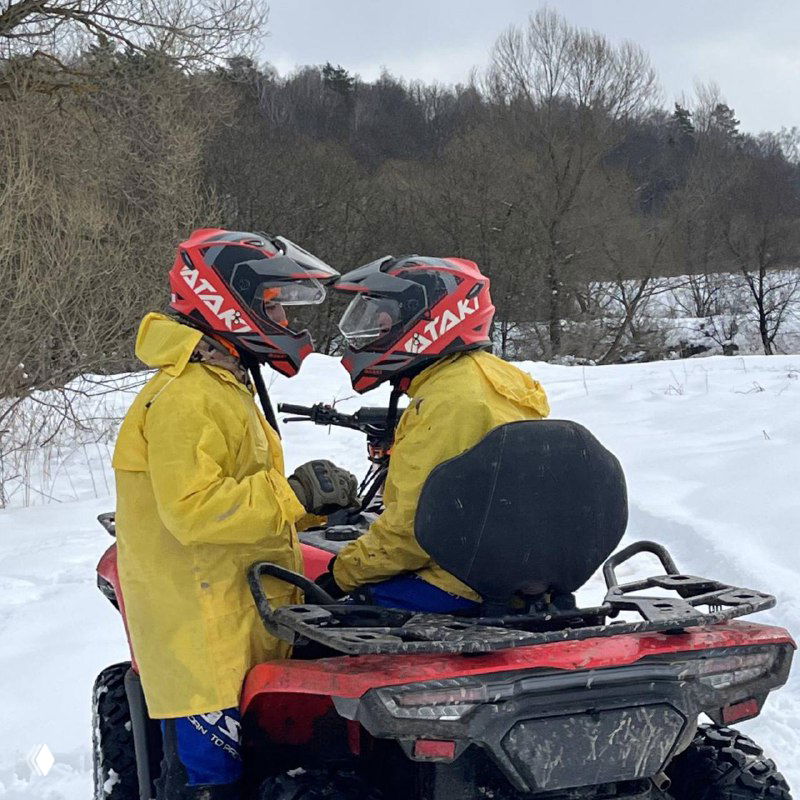 Два участника в ярких дождевиках и шлемах сидят лицом друг к другу на квадроцикле на заснеженном участке пути — зима в Подмосковье.
