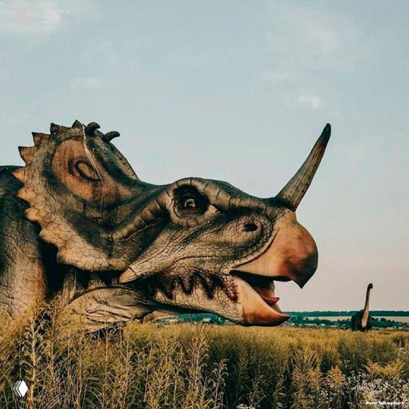 Крупный план головы стилизованной скульптуры динозавра на фоне поля и неба — заброшенный динопарк на территории туристического комплекса «Золотой город», Тульская область.