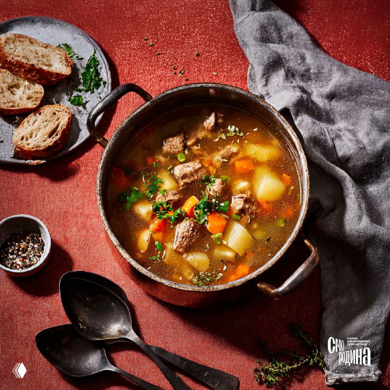 Чугунный казан с наваристым супом лилэпс, рядом ломти хрустящего домашнего хлеба и столовые ложки на красном фоне, сервировка для подачи.