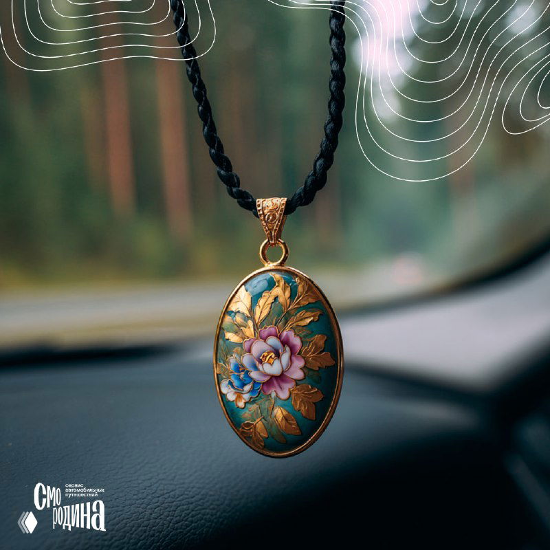 Овальная подвеска-финифть с цветочным узором и золотой окантовкой, крупный план на фоне размытых деревьев за лобовым стеклом автомобиля.