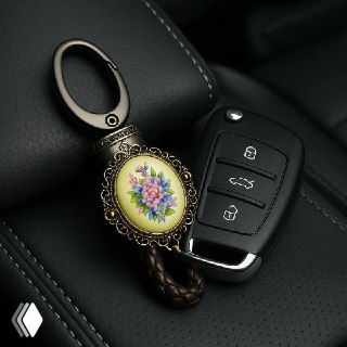 Ключи от автомобиля с брелоком-финифтью, брелок с миниатюрной цветочной росписью лежит на чёрной кожаной поверхности сиденья или подлокотника.