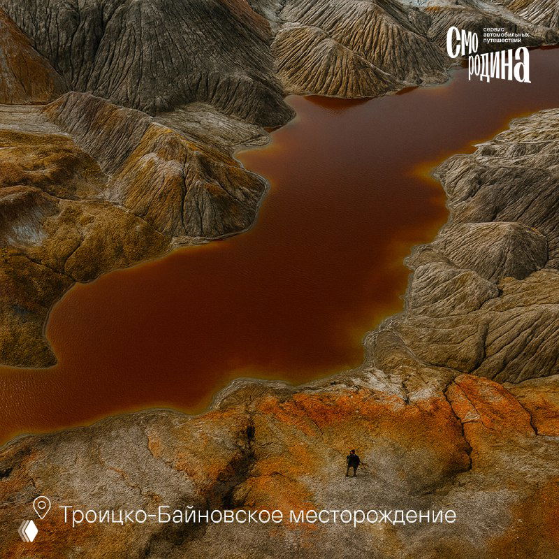 Пейзаж с марсианскими оттенками: извилистое озеро и неровные грунтовые холмы с красноватыми и коричневыми породами вдалеке.