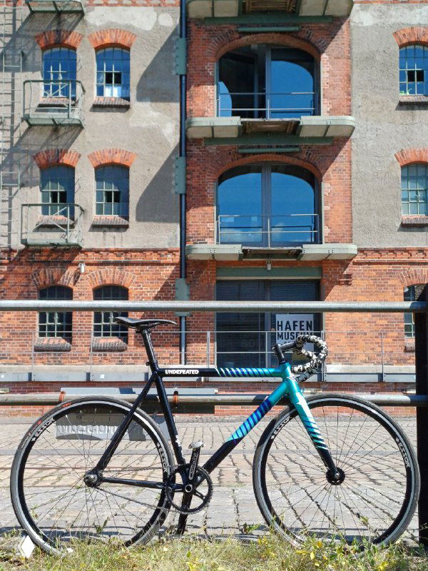 Фикс велосипед State Cycles Undefeated припаркован у кирпичного фасада порта в Бремене — видно профиль рамы, колёса и контраст городской текстуры.