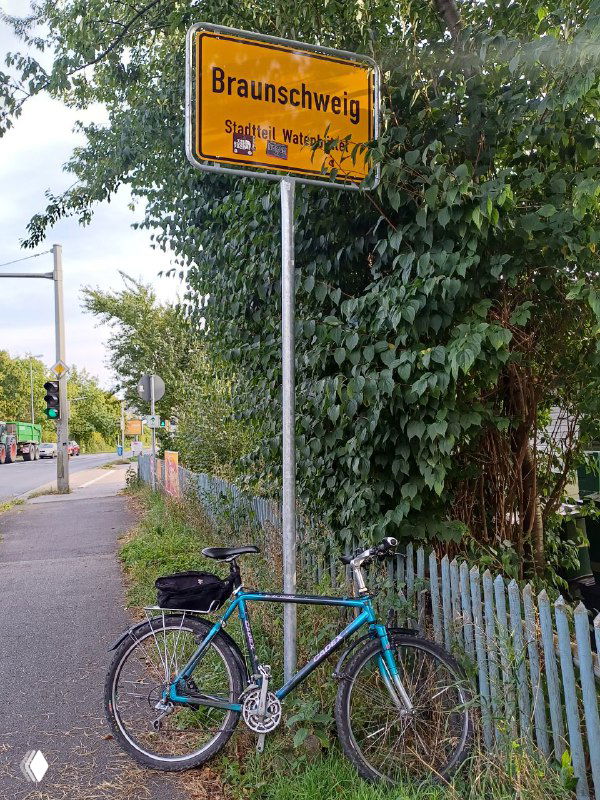 Голубой байк припаркован у дорожного знака «Braunschweig» на фоне зелёных кустов и деревянного забора, сельская сцена и тротуар.