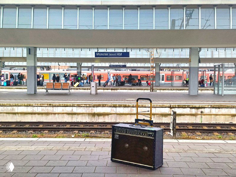 Гитарный комбо Roland на платформе железнодорожной станции в Германии — композиция подчёркивает тему перевозок и доставки музыкального оборудования между странами.