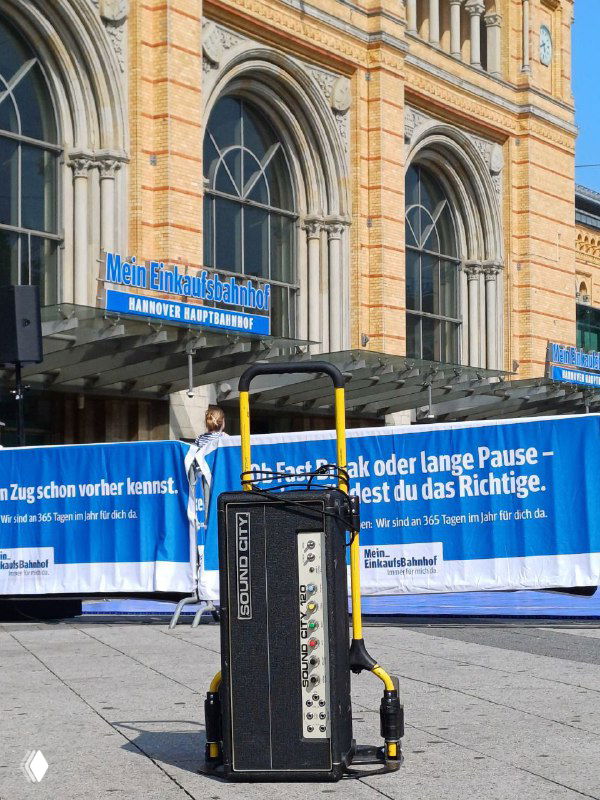 Усилитель Sound City на колёсной стойке перед зданием в Ганновере — видны синий рекламный баннер, фасад вокзала и городской фон, указывающий на место покупки.