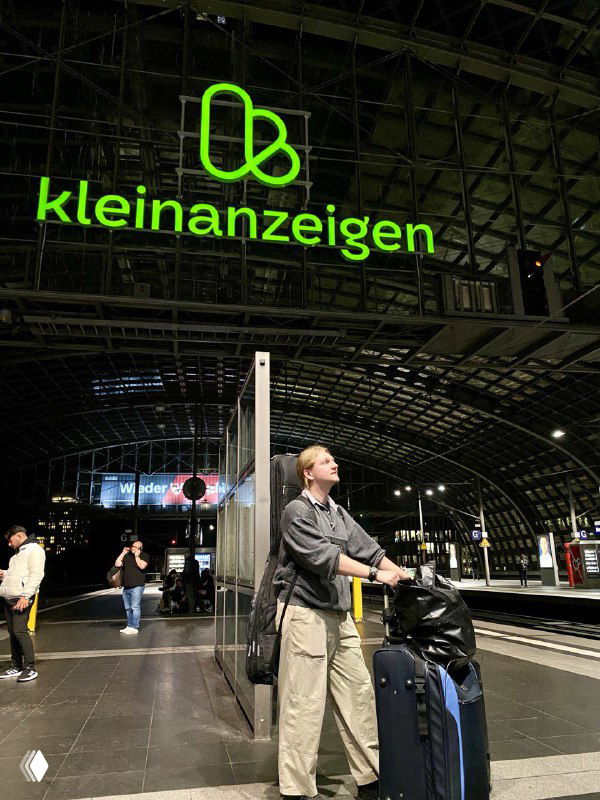 Пассажир с чемоданом и рюкзаком на ночной платформе на фоне зелёного логотипа kleinanzeigen; образ охотника за вещами и поездок ради выкупа.