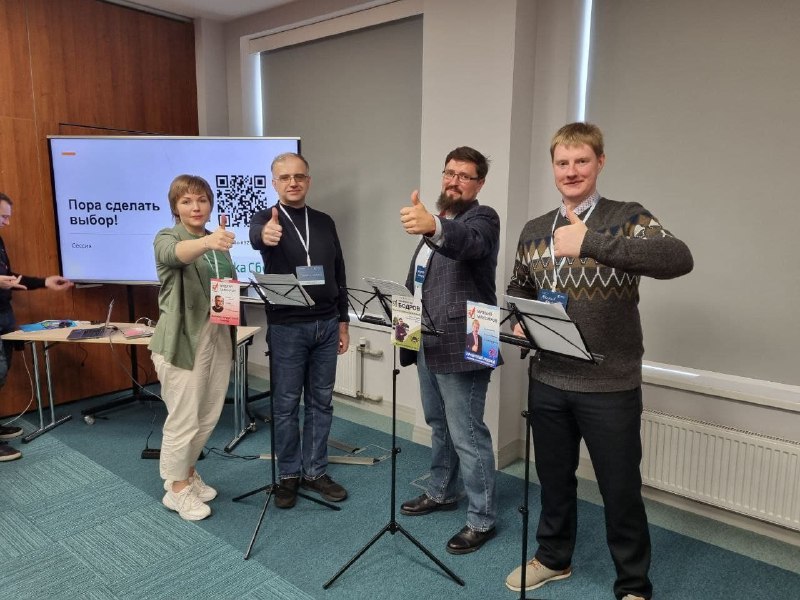 Четверо людей в конференц-зале у микрофонов: участники дебатов позируют вместе, видна сцена, экран и экипировка