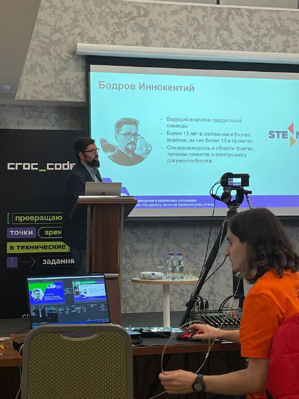 Докладчик стоит за трибуной на сцене конференции Analyst Days; виден экран со слайдом, камера и оператор на переднем плане.
