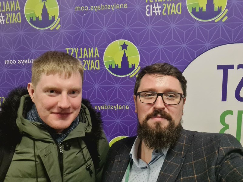 Селфи двух мужчин на фоне баннера конференции Analyst Days: видны логотипы мероприятия, фиолетовая заставка и улыбающиеся участники.