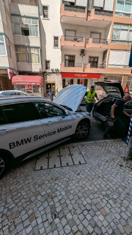 Уличная сцена с автомобилем BMW Service Mobile: открытый капот, техник у передней части машины, видна городская парковка и пешеходная зона.