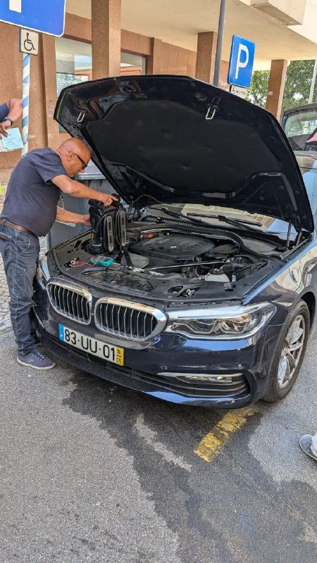 Крупный план мотора BMW с поднятым капотом, техник работает с бустером и клеммами, вокруг видна городская застройка и разметка парковки.