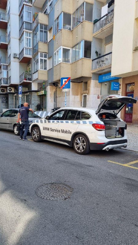Общий вид: BMW Service Mobile с открытым багажником, техник рядом, автомобиль припаркован на узкой односторонней улице у жилого дома.