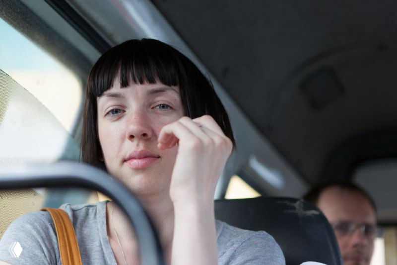 Человек в автомобиле смотрит в окно, слегка тронутый эмоциями, на заднем плане пассажир, ощущение пути, размышлений и усталости.