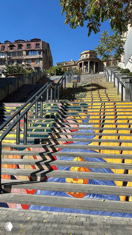 Городская лестница на улице с яркой росписью ступеней, металлическими перилами и зелёным деревом на фоне синего неба.