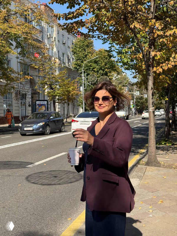 Женщина в солнцезащитных очках держит бумажный стакан кофе и улыбается на городской улице среди осенних деревьев и автомобилей