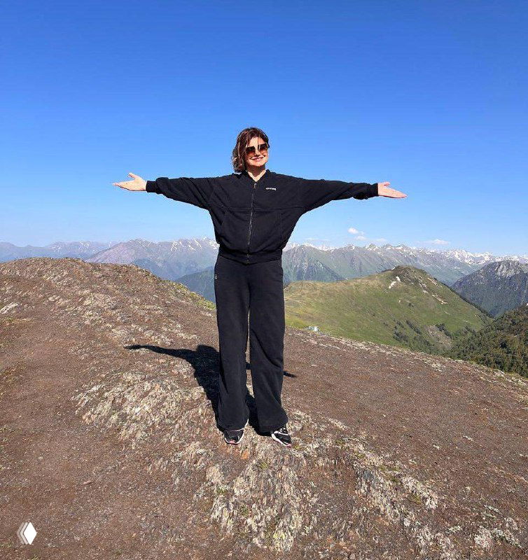 Женщина в тёмной одежде стоит с расправленными руками на скалистом хребте, вокруг горы и голубое небо, образ дистанции и спокойствия.