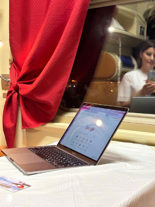 Ноутбук с открытым браузером на столике в купе поезда, красная штора слева и зеркальное отражение пассажирки.