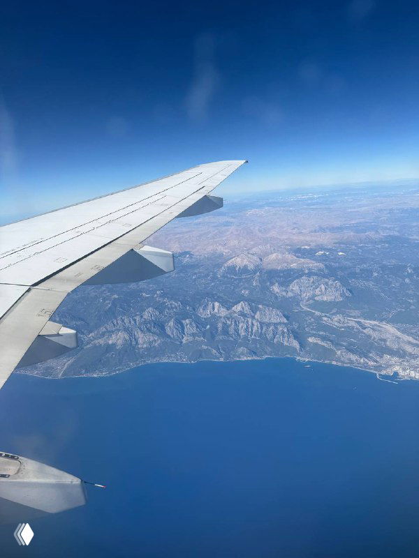 Вид из иллюминатора самолёта: крыло в левом углу, под ним побережье и горы у моря под чистым голубым небом.