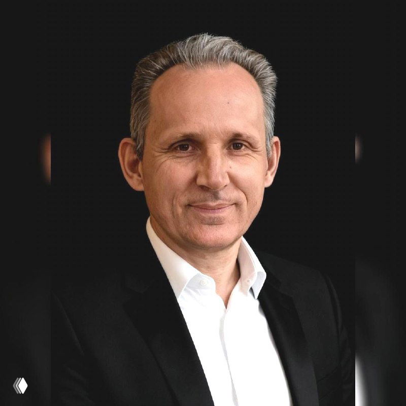 Фотография портретного плана: взрослый мужчина в пиджаке и белой рубашке на тёмном фоне, деловой нейтральный вид, смотрит в камеру.