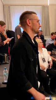 Панорамный кадр с участниками форума в Тюмени: в зале люди слушают доклады, мужчина в профиль, на столах бутылки воды и бейджи.