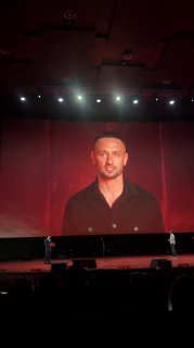 Широкий план сцены с экраном, на котором демонстрируется видео с крупным планом человека — кадр во время выступления или трансляции на большом экранe.