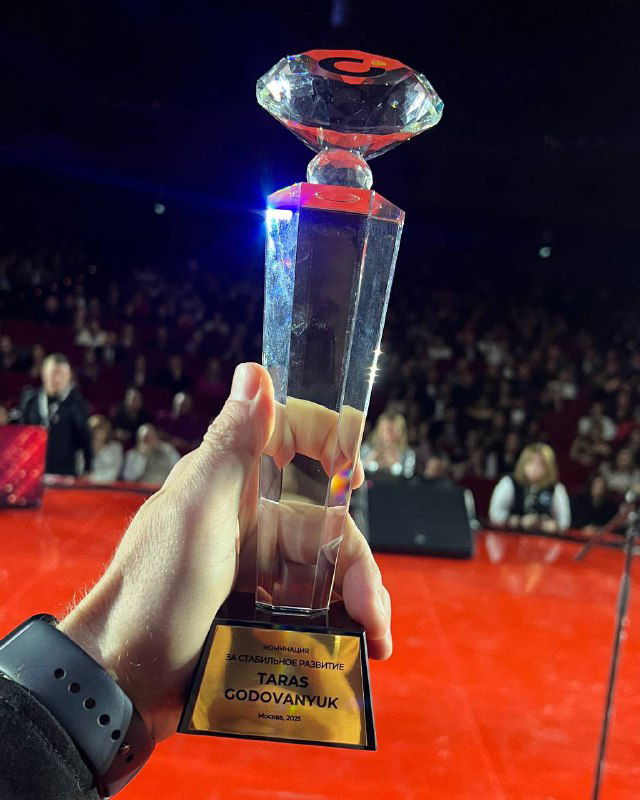 Рука держит прозрачную статуэтку-приз на фоне заполненного зала — момент награждения и символ признания участников мероприятия «Итоги 2024 UDS».