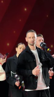 Человек в чёрной куртке на сцене держит награду, крупный план во время церемонии — спикер или лауреат вечера на фоне красной подсветки.