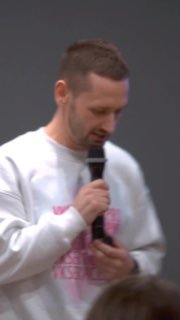 Вертикальный кадр: мужчина держит микрофон в белом свитшоте с розовым принтом на деловом выступлении или митапе; фон и публика размыты