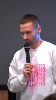 Человек в белом свитшоте с розовым принтом держит микрофон; вертикальный портрет на нейтральном фоне как иллюстрация формата видео.