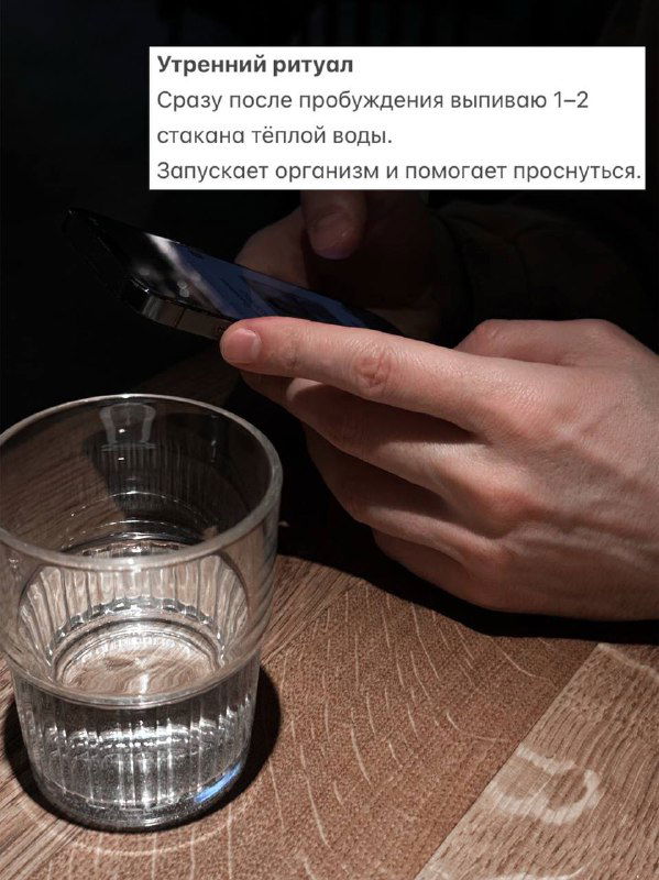 Крупный план руки и стакана тёплой воды на столе; фотография иллюстрирует утренний ритуал — питьё воды сразу после пробуждения для бодрости и запуска организма.