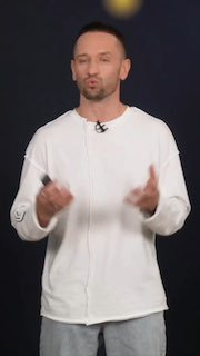 Ведущий в белой толстовке на тёмной сцене во время презентации: жестикуляция руками, микрофон и атмосферный фон — промо‑кадр онлайн‑трансляции UDS