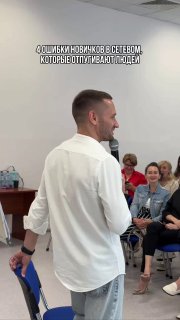 Мужчина-спикер в белой рубашке держит микрофон и обращается к аудитории на встрече по сетевому маркетингу; слушатели сидят и улыбаются.