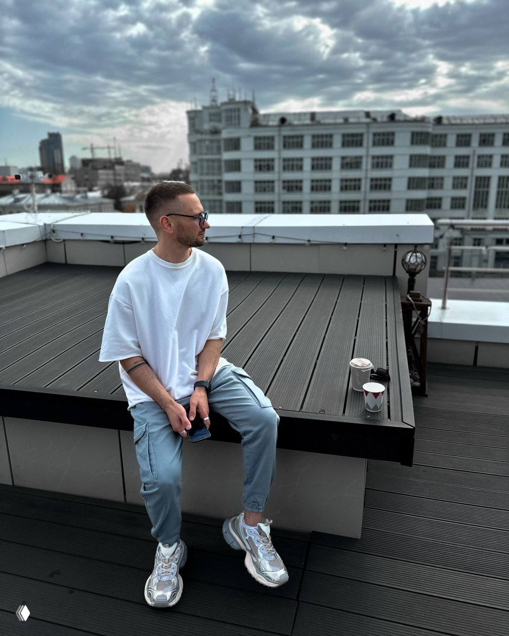 Мужчина сидит на крыше здания, смотрит в сторону города; городской пейзаж и чай в стакане рядом создают неформальную атмосферу размышлений.
