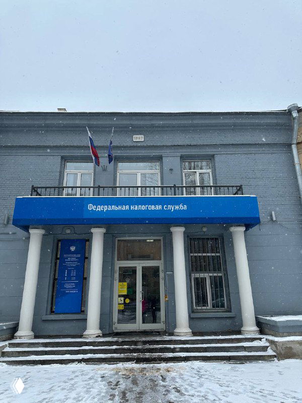 Здание Федеральной налоговой службы зимой: вход с колоннами и синим козырьком с надписью, российские флаги на фасаде и снежная дорожка перед входом