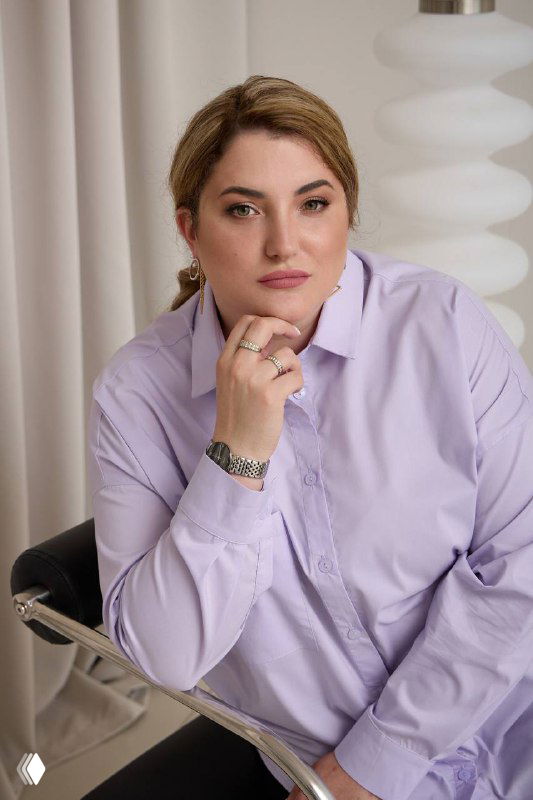 Фотография женщины в светлой рубашке, сидящей в кресле на фоне нейтрального офисного интерьера; иллюстрация к теме.