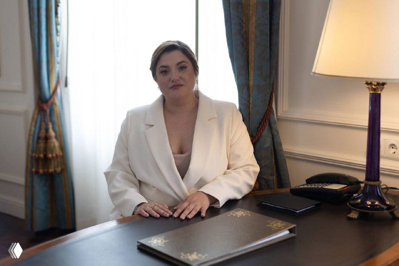 Деловой портрет женщины в кабинете: сидит за столом в белом пиджаке, на столе ноутбук, лампа и деловые папки — иллюстрация к новости.