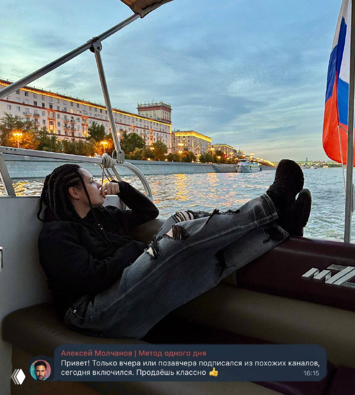 Человек отдыхает, лежа на борту прогулочного катера; на заднем плане видна набережная с вечерними огнями и российский флаг на носу судна.