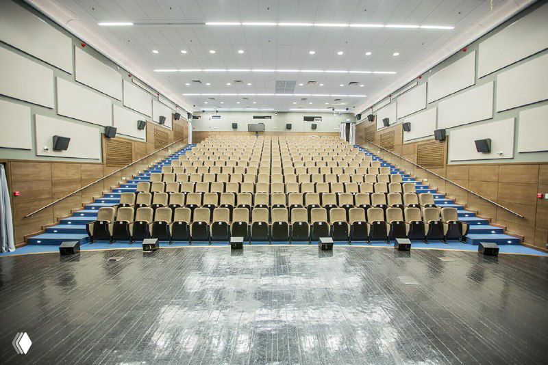 Фотография пустого зрительного зала примерно на 300 мест: вид с сцены, аккуратные ряды кресел, деревянный пол, белые стены и сцена с оборудованием.