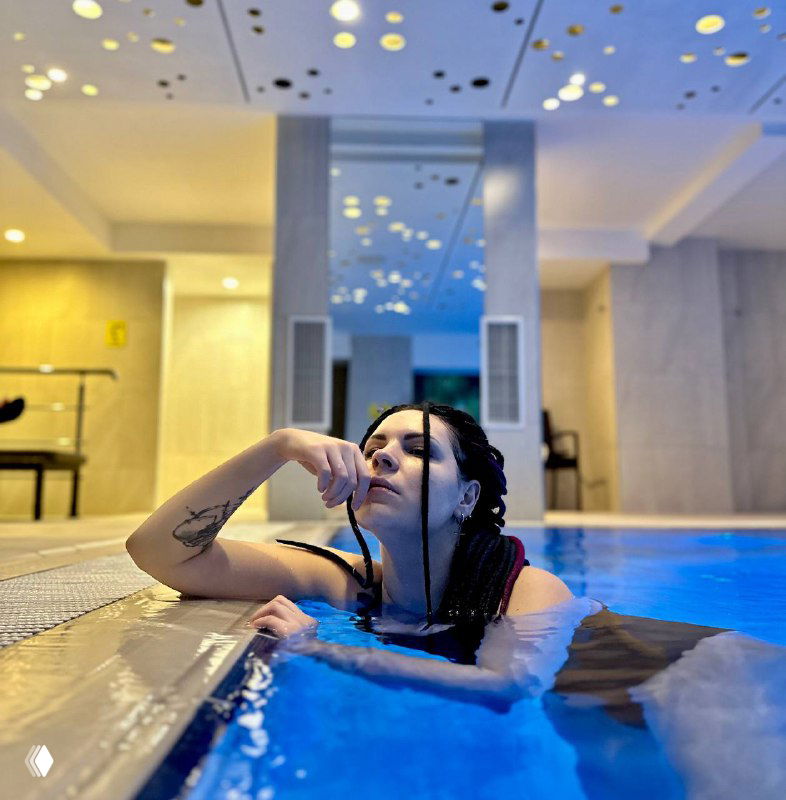 Женщина с заплетёнными волосами в бассейне внутреннего спа, опирается на бортик; видны татуировки, расслабленная задумчивая поза автора.