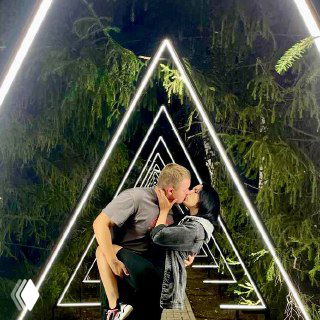 Пара целуется внутри светящейся инсталляции из треугольных рам на фоне темных хвойных деревьев — ночная романтическая сцена.