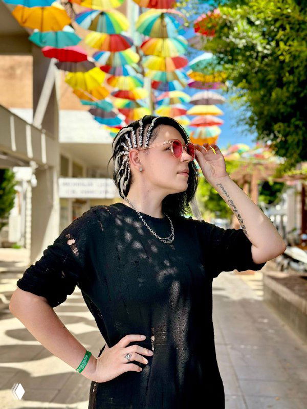 Автор поста в чёрной футболке и солнцезащитных очках с плетёными косами, стоит на улице под рядом ярких подвесных разноцветных зонтиков, поза уверенная.