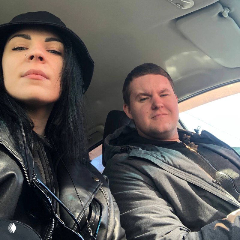 Селфи двух людей в салоне автомобиля: женщина в черной панаме и мужчина в куртках смотрят в камеру, дневной свет, повседневный контекст.