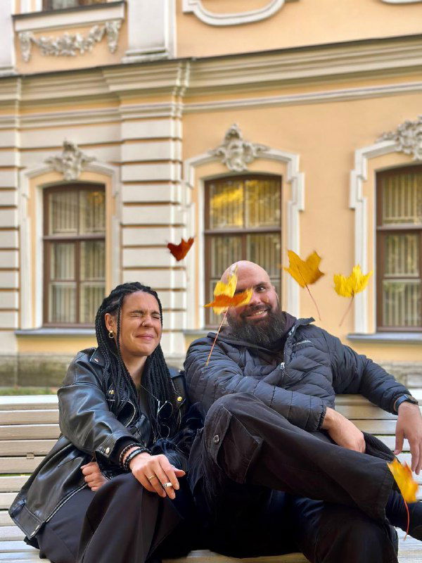 Двое людей в тёплой одежде сидят на скамейке перед историческим зданием, улыбаются; осенние листья вокруг, городской фон.