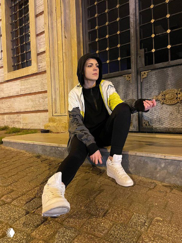 Женщина в худи и кроссовках сидит на тротуаре у декоративной двери ночью в городском пространстве, расслабленная поза и уличная атмосфера.