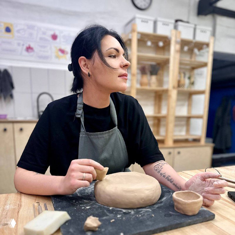 Портрет в гончарной мастерской: женщина лепит миску из глины на столе, вокруг полки с керамикой и инструменты на фоне мастерской.