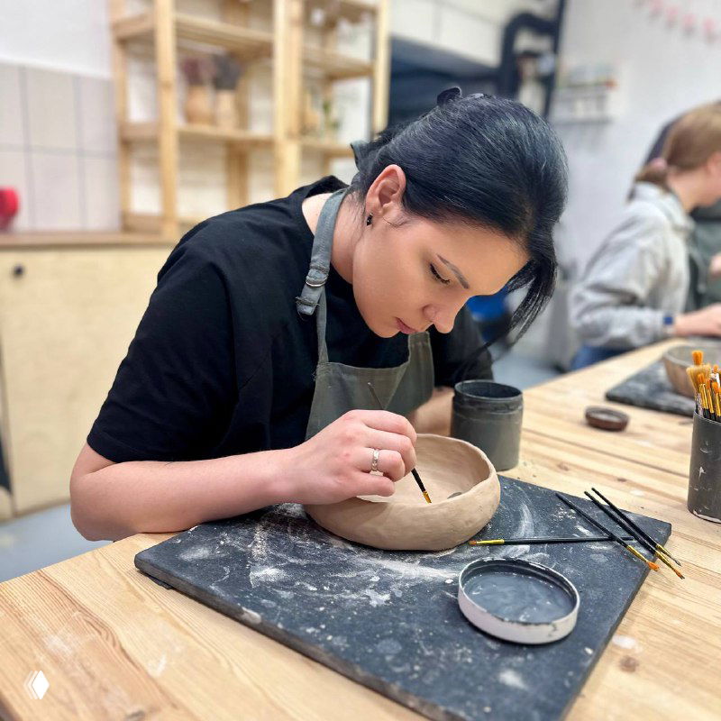 Крупный план: женщина сосредоточенно обрабатывает керамическую чашу на рабочем столе, вокруг материалы и кисточки для декора.