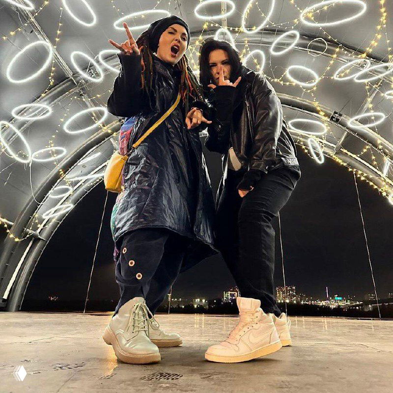 Две молодые женщины в тёплой одежде позируют ночью под аркой со светящимися кольцами; снимок в полный рост на городской площадке.