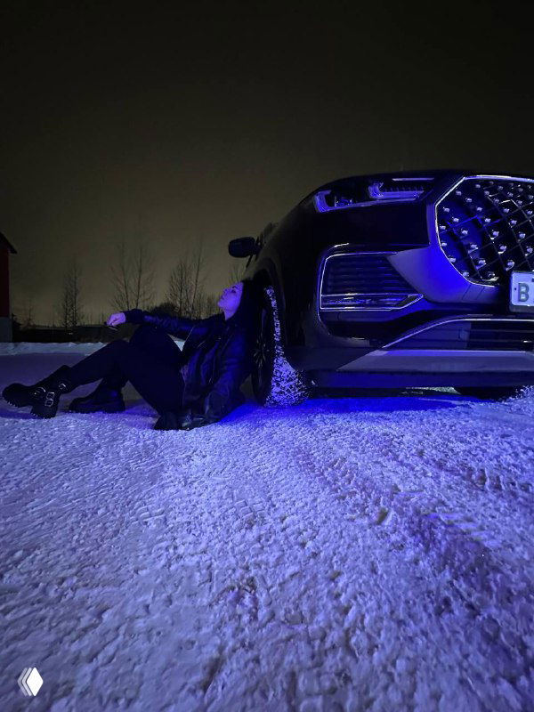 Ночной кадр спереди: премиальный внедорожник с подсветкой, автор сидит у бампера на снегу — сцена для фото о путешествиях и достатке.