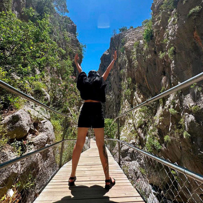 Человек стоит спиной на подвесном мосту между скал в лучах солнца, подняв руки вверх — символ свободы и преодоления